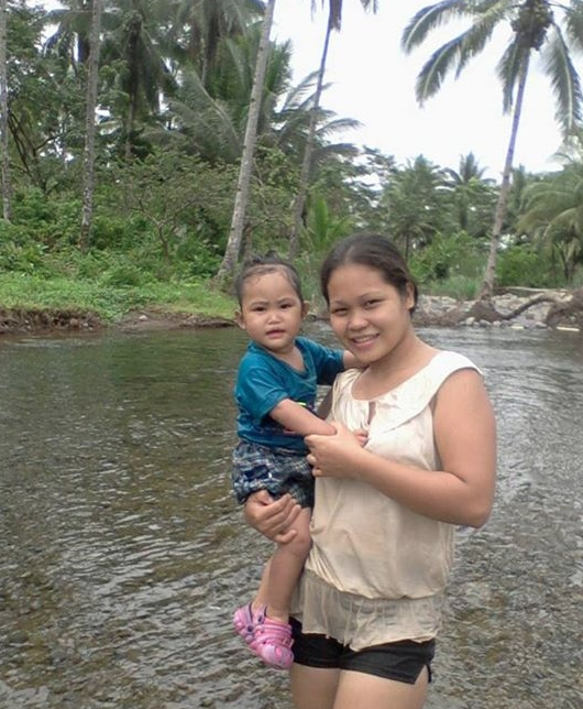 A young woman holding a toddler while standing in a shallow river among palm trees in the Philippines